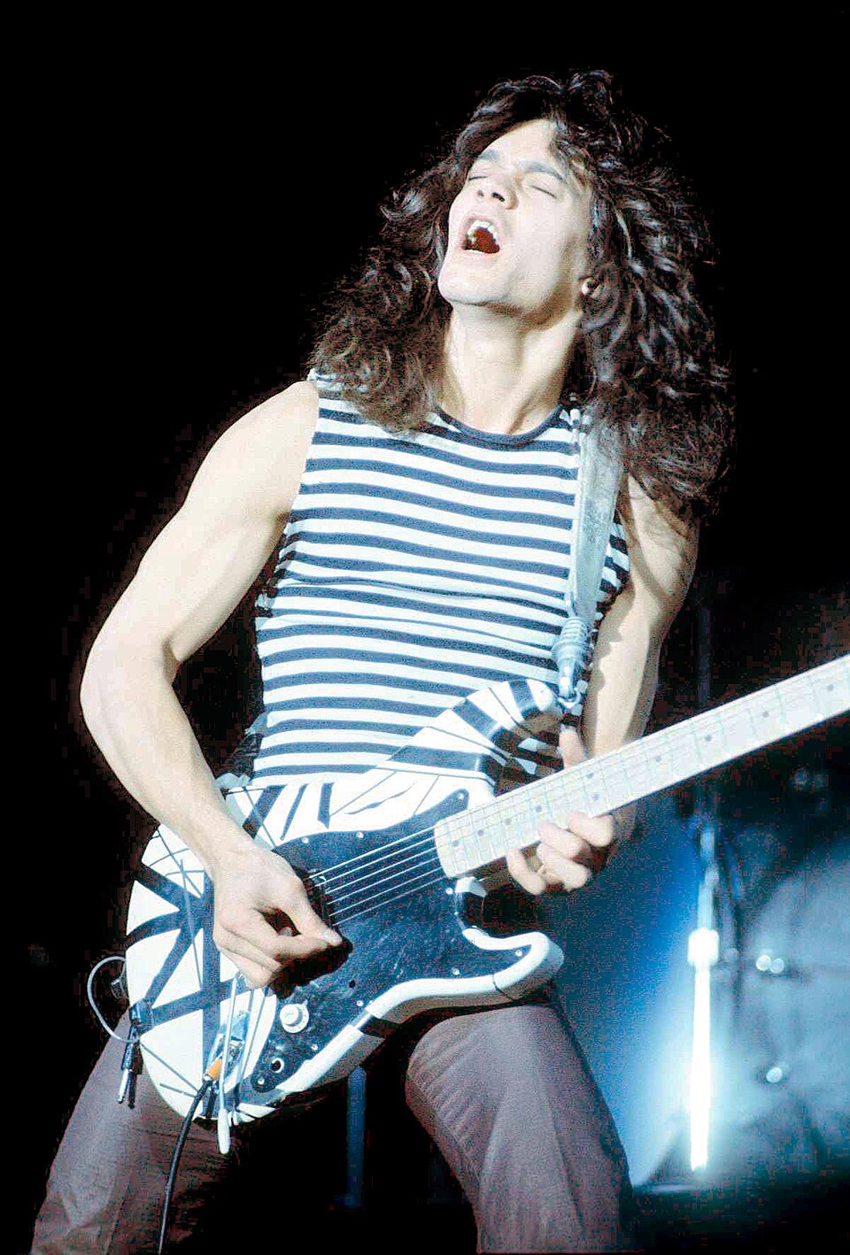 Male guitarist in a blue-and-white striped tank top passionately playing an electric guitar on stage, eyes closed.